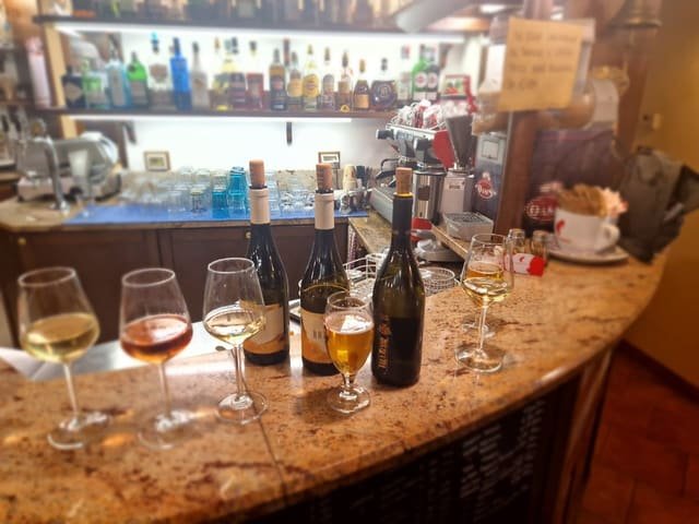 Cinque calici di vino bianco, ambrato e rosato allineati sul bancone di marmo con tre bottiglie aperte alle spalle