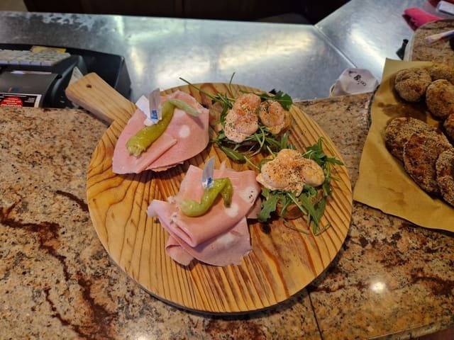Tagliere tondo di legno con crostini di mortadella e peperoncino verde e crostini con gamberi e rucola, appoggiato sul piano di marmo del bancone