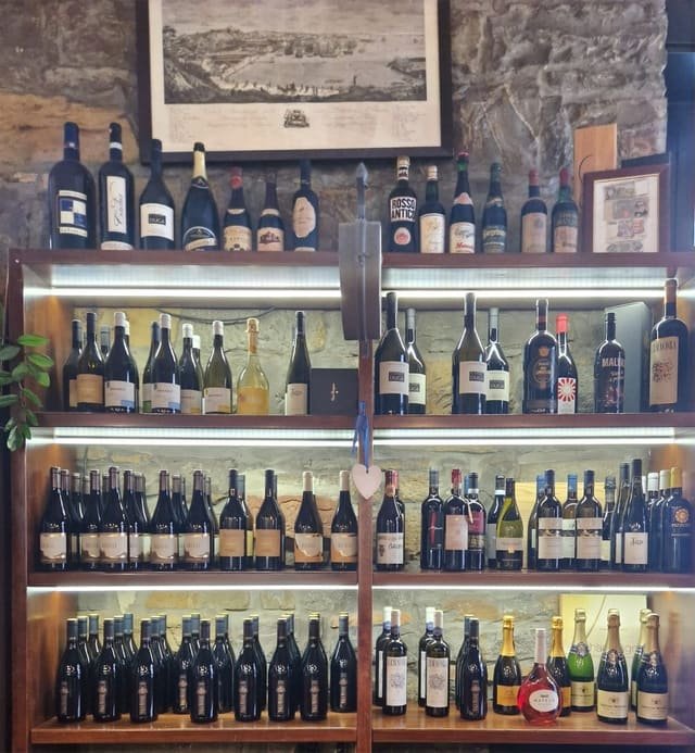 Scaffalature di legno illuminate contro il muro di pietra, piene di bottiglie di vino friulano, con una stampa antica del porto di Trieste appesa sopra
