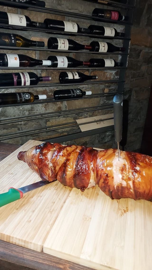 Porchetta intera dorata appoggiata su un tagliere di legno chiaro, con il coltello e il forchettone, davanti alla rastrelliera dei vini sul muro di pietra