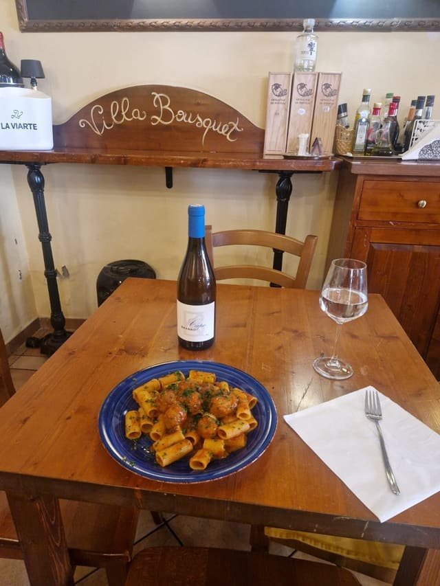 Rigatoni con polpette al sugo su un piatto blu, accanto una bottiglia di bianco del Collio e un calice, sul tavolo di legno della saletta